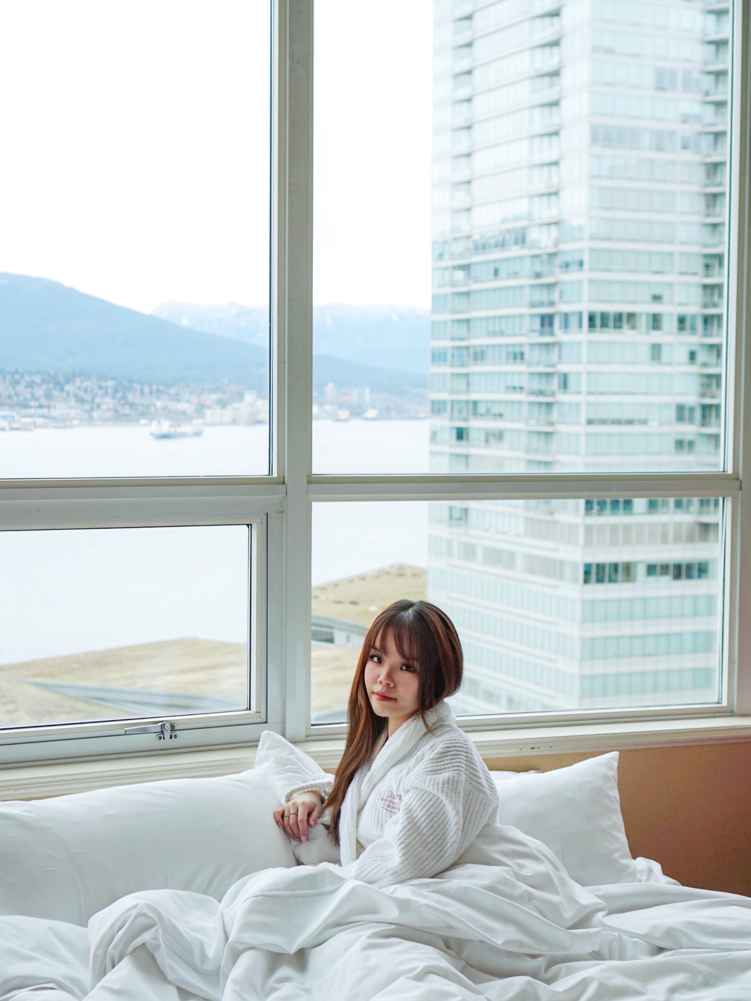 Girl sitting in a white fluffy bed with white pillows overlooking Vancouver's coal harbour in Vancouver Marriott Pinnacle